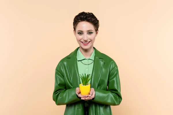 Happy woman in green jacket holding plant in yellow flowerpot isolated on beige — Stock Photo