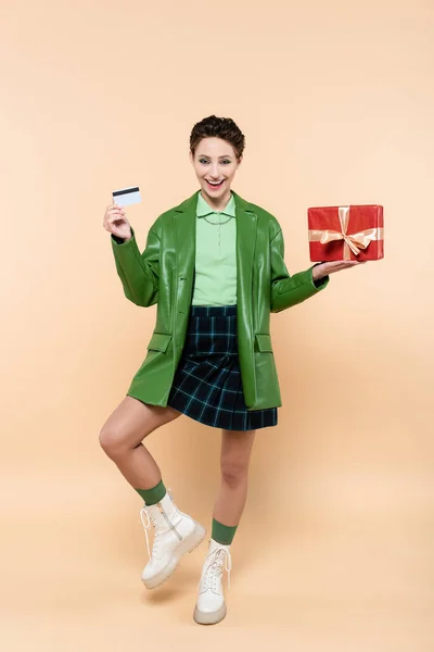 Vista completa de la mujer alegre en chaqueta verde y falda a cuadros posando con tarjeta de crédito y caja de regalo en beige - foto de stock
