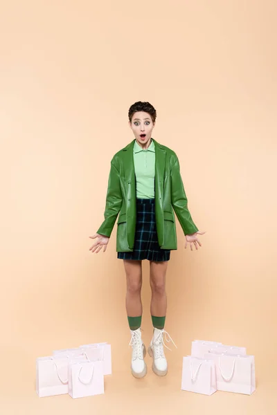 Astonished woman in green jacket, checkered skirt and white boots levitating near shopping bags on beige — Foto stock