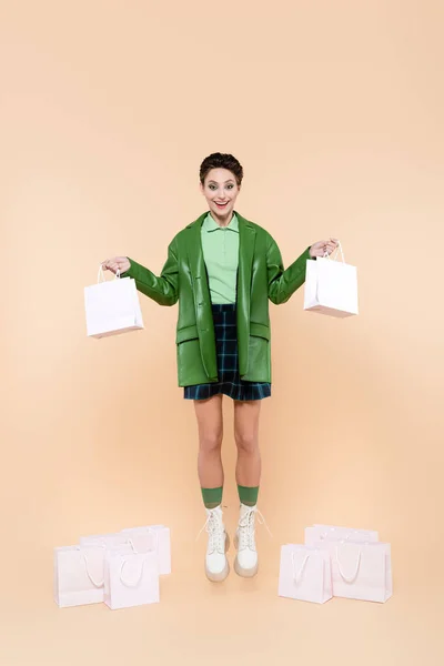 Full length view of excited woman in trendy clothes levitating with shopping bags on beige — стоковое фото