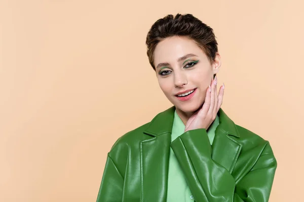 Young brunette woman in green leather jacket touching face and smiling at camera isolated on beige — Foto stock