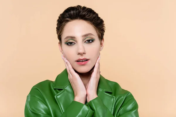 Brunette woman in green leather jacket touching face while looking at camera isolated on beige — Stockfoto