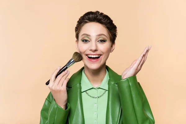 Excited woman applying face powder with cosmetic brush and pointing with hand isolated on beige — Stock Photo