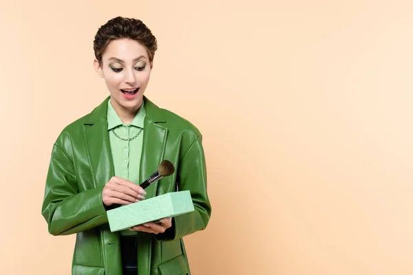 Amazed woman in stylish jacket holding gift box and cosmetic brush isolated on beige — Stock Photo