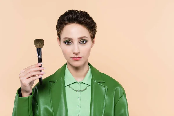 Brunette woman in green leather jacket holding cosmetic brush isolated on beige — Stock Photo