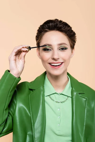 Happy and stylish woman applying mascara while looking at camera isolated on beige — Stock Photo