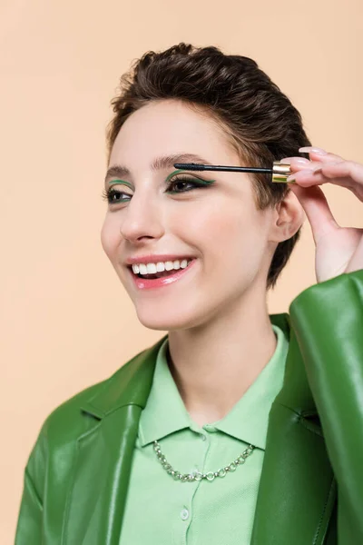 Mujer morena complacida con pelo corto aplicando rímel aislado en beige - foto de stock