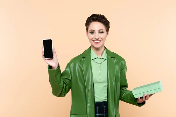 Allegra donna bruna con scatola regalo che mostra smartphone con schermo bianco isolato sul beige — Foto stock
