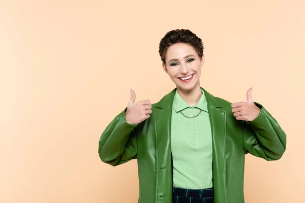 Joyful woman in green stylish jacket showing thumbs up isolated on beige — стоковое фото