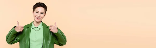 Cheerful young woman in green trendy jacket showing thumbs up isolated on beige, banner — Stockfoto