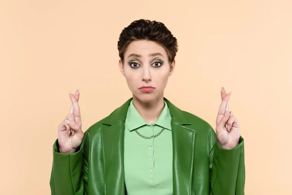 Discouraged woman in green leather jacket holding crossed fingers isolated on beige — стоковое фото