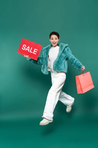 Mujer excitada en chaqueta de piel sintética y pantalones corriendo con tarjeta de venta y bolsa de compras en verde - foto de stock
