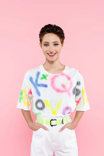 Joyful woman smiling at camera while posing with hands in pockets isolated on pink — Foto stock