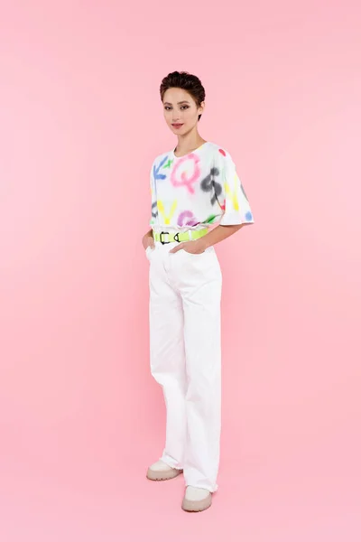 Full length view of stylish smiling woman standing with hands in pockets of white trousers on pink — Stockfoto