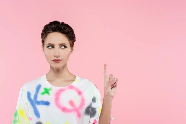 Seria morena mujer en elegante camiseta apuntando hacia arriba con el dedo aislado en rosa - foto de stock