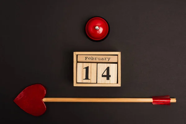 Вид стрелы в форме сердца рядом с кубиками с 14 февраля надписью и колоколом на черном — стоковое фото