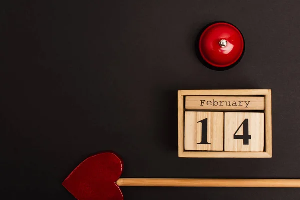 Top view of heart-shaped arrow near wooden cubes with 14 february lettering and bell on black — Fotografia de Stock