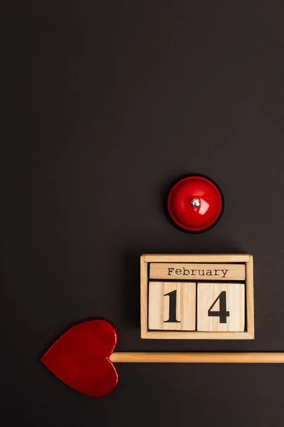 Top view of heart-shaped arrow near wooden cubes with 14 february lettering and metallic bell on black — Stock Photo