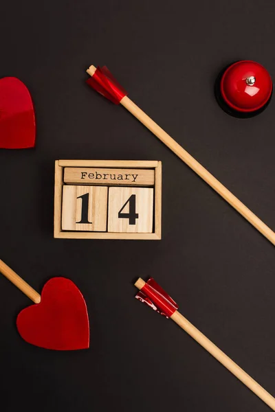 Top view of heart-shaped arrows near cubes with 14 february lettering and bell on black — Fotografia de Stock