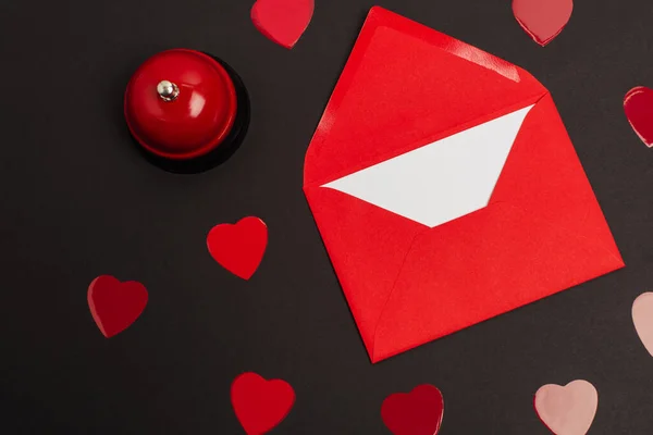 Vista superior de sobre rojo con carta de amor y corazones cortados de papel cerca de campana aislada en negro - foto de stock