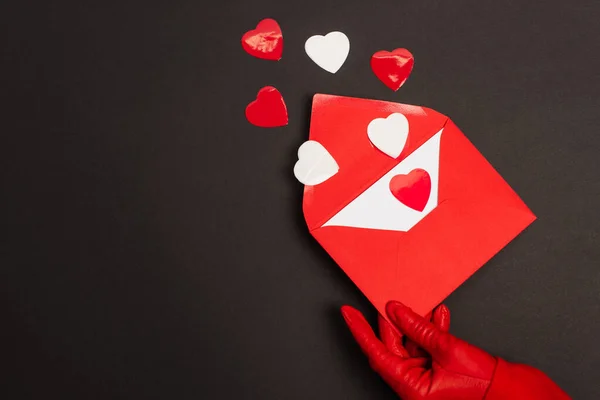 Cropped view of person in red glove holding envelope with love letter and paper cut hearts on black — Stock Photo