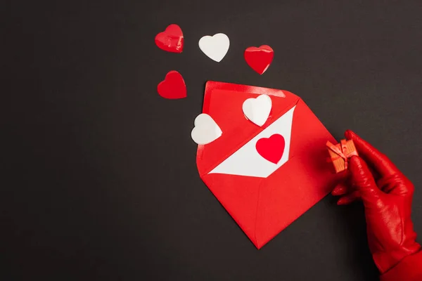 Vista recortada de la persona en guante rojo sosteniendo una pequeña caja de regalo cerca del sobre con carta de amor y corazones cortados en papel en negro - foto de stock