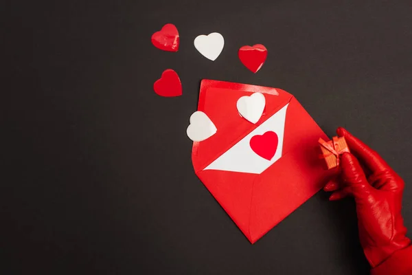 Cropped view of person in red glove holding tiny gift box near envelope with love letter and paper cut hearts on black — Stock Photo