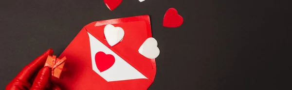 Cropped view of person in red glove holding tiny gift box near envelope and paper cut hearts isolated on black, banner — Foto stock