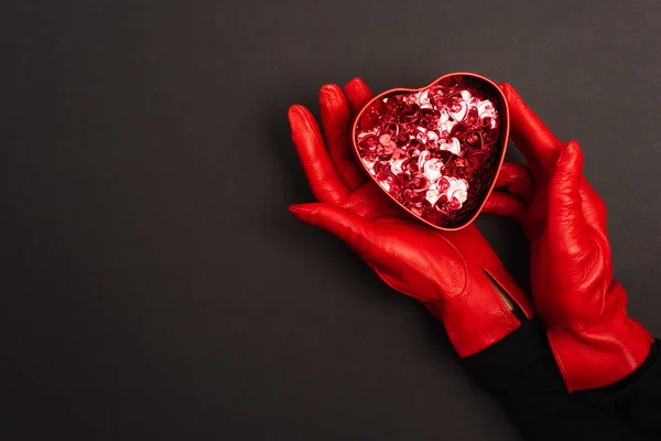 Cropped view of woman in red gloves holding metallic heart-shaped box on black — Stock Photo