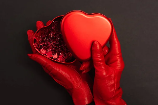Vista superior da mulher em luvas vermelhas segurando caixa metálica em forma de coração no preto — Fotografia de Stock
