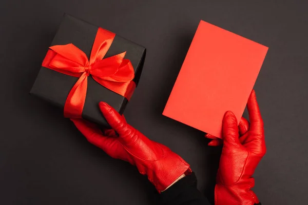 Vista recortada de la persona en guantes rojos sosteniendo envuelto caja de regalo y sobre en negro - foto de stock