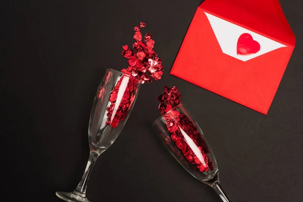 Top view of champagne glasses with red confetti hearts near envelope on black — Stock Photo
