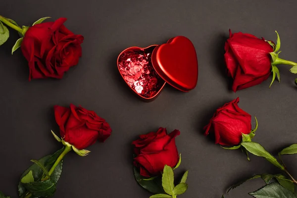 Top view of metallic box with red confetti hearts near roses on black — Stock Photo