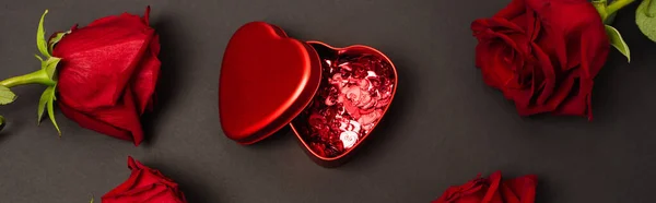 Vue du dessus de la boîte métallique avec des cœurs de confettis rouges près des roses sur noir, bannière — Photo de stock