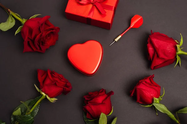 Disposición plana de caja metálica en forma de corazón cerca de regalo envuelto, oscuro y rosas, en negro - foto de stock