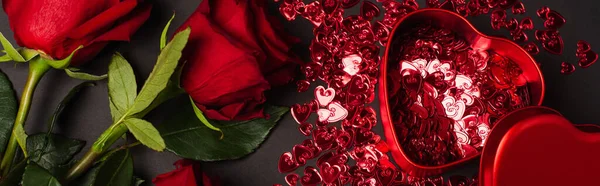 Top view of red roses near metallic heart-shaped box and shiny confetti on black, banner — Foto stock