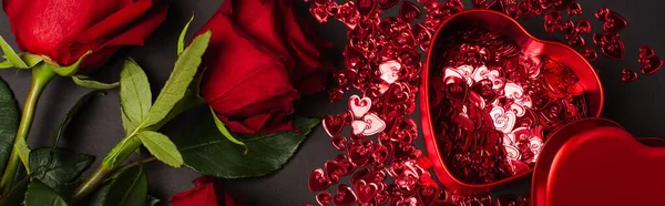 Vista dall'alto di rose rosse vicino a scatola metallica a forma di cuore e coriandoli lucidi su nero, banner — Foto stock