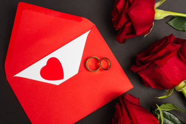 Top view of red roses near envelope and golden rings on black — Foto stock