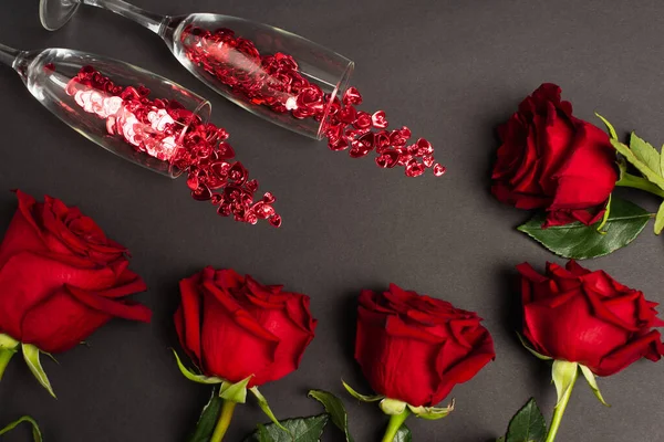 Top view of red roses near glasses with confetti on black — Stock Photo