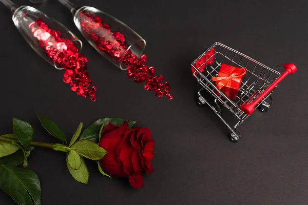 Top view of red rose near glasses with confetti and tiny present in shopping cart on black — Stock Photo