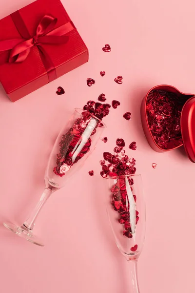 Top view of champagne glasses with confetti hearts near metallic heart-shaped box and present on pink — Stock Photo