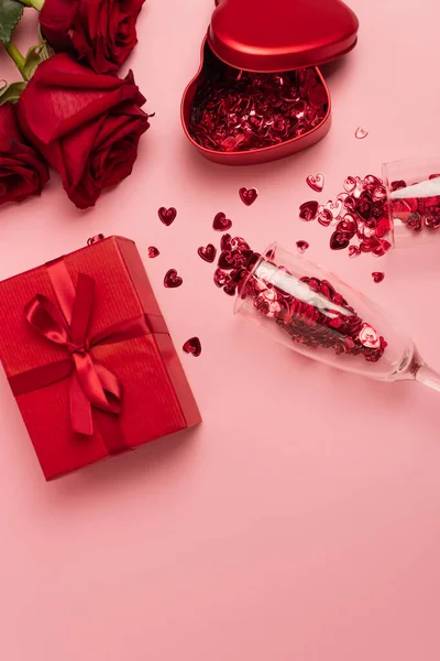 Vista dall'alto di bicchieri di champagne con cuori di coriandoli vicino a scatole regalo e rose rosse su rosa — Foto stock