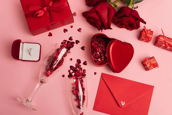 Top view of champagne glasses with shiny confetti near gift boxes, engagement ring and red roses on pink — Foto stock