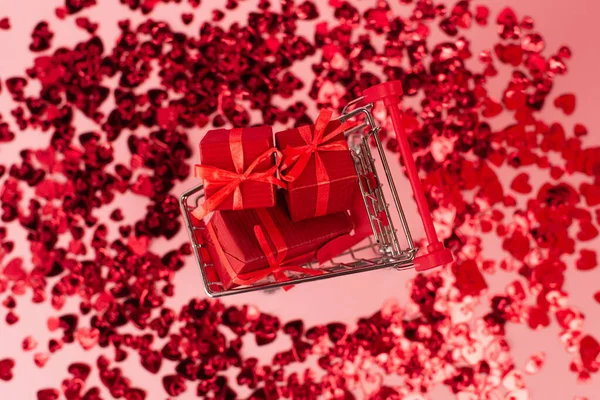 Top view of small shopping cart with presents near shiny red confetti hearts on pink — Fotografia de Stock