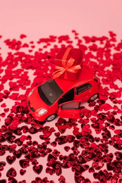 Високий кут зору іграшкового автомобіля з загорнутим подарунком біля блискучих червоних сердець конфетті на рожевому — стокове фото