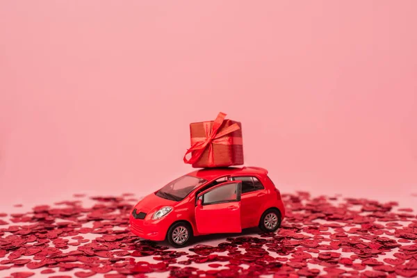 Toy car with wrapped present near shiny red confetti hearts isolated on pink — Stock Photo
