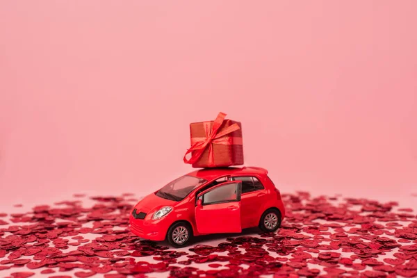 Voiture jouet avec enveloppé présent près de cœurs confettis rouge brillant isolé sur rose — Photo de stock