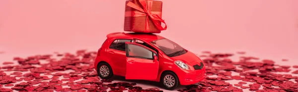 Spielzeugauto mit eingewickeltem Geschenk in der Nähe leuchtend roter Konfetti-Herzen isoliert auf rosa, Banner — Stockfoto