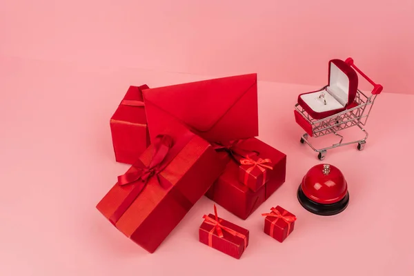 High angle view of red wrapped gift boxes and envelope near shopping cart with engagement ring on pink — Foto stock