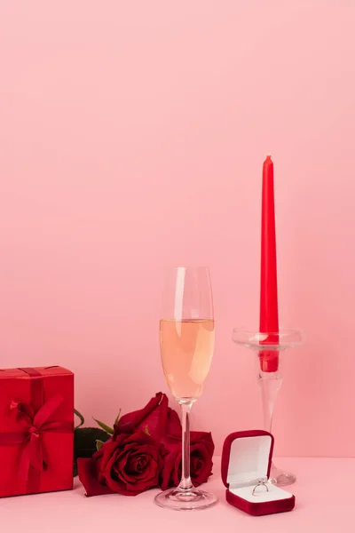 Coupe de champagne près boîte cadeau, roses, bague de fiançailles et bougie sur rose — Photo de stock